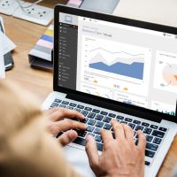 Person arbeitet an einem Laptop mit Diagrammen und Daten, eine zweite Person gestikuliert daneben.