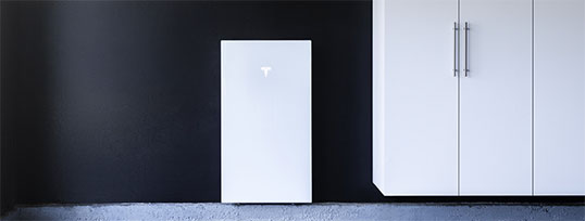Weiße Tesla Powerwall an schwarzer Wand montiert, kompakt neben einem weißen Hängeschrank platziert.